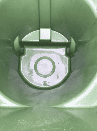 Close-up view of the inside of a green plastic trash bin, showing its circular base with a central ring pattern and hinge mechanism. The surface appears slightly worn and reflective, ideal for before-and-after shots by a trash can cleaning service.