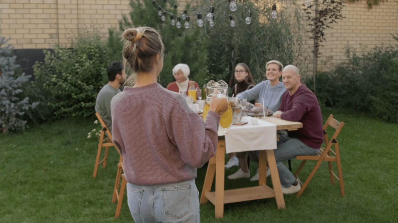 A woman with her hair in a bun stands holding a jug of juice, facing a wooden table outdoors. Five people, both young and old, smile and look at her. They sit in a green garden with string lights and plants, gathered for a meal.