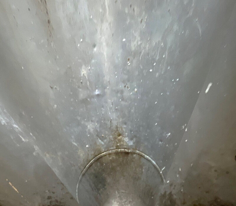 A close-up photo of a metal pipe or tunnel, viewed from inside. The surface is shiny, with grime and water stains, and there is a circular ridge near the center-bottom. The lighting highlights texture, creating a reflective and slightly dirty appearance.