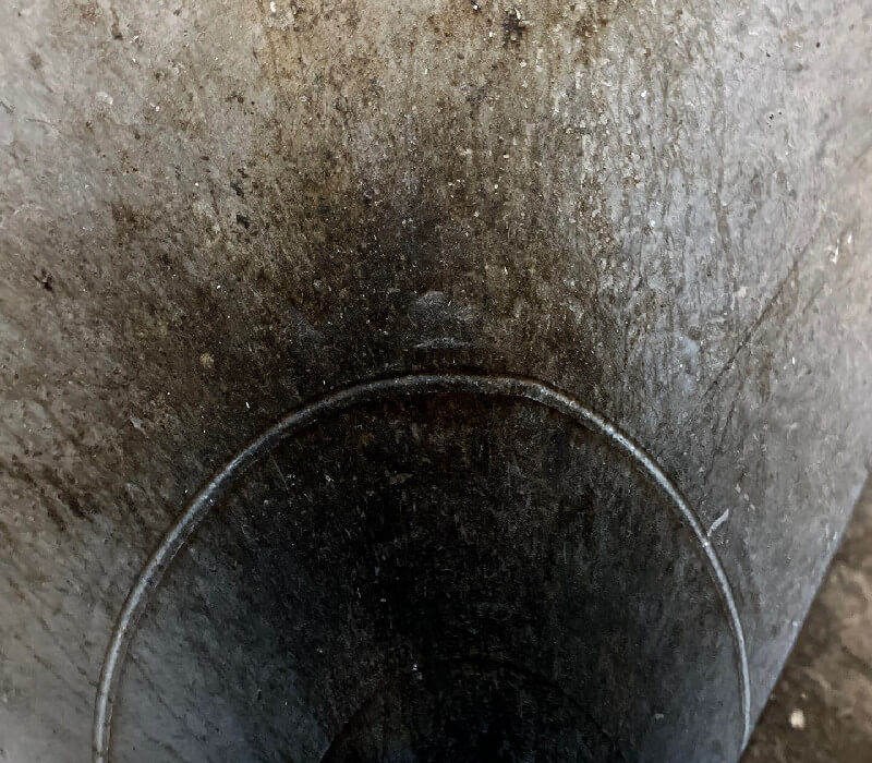 A close-up view looking down into a dark, cylindrical, concrete or metal well or pipe, with textured, stained surfaces and a circular rim. The interior fades into darkness, and dirt or grime is visible along the walls.