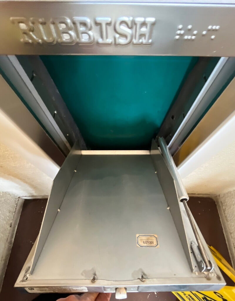 A close-up of a metal rubbish chute with the word "RUBBISH" embossed at the top. The chute is open, revealing a hinged tray labeled "ROTATION" and green interior walls. The surrounding frame is metallic, and part of a yellow sign is visible at the bottom.