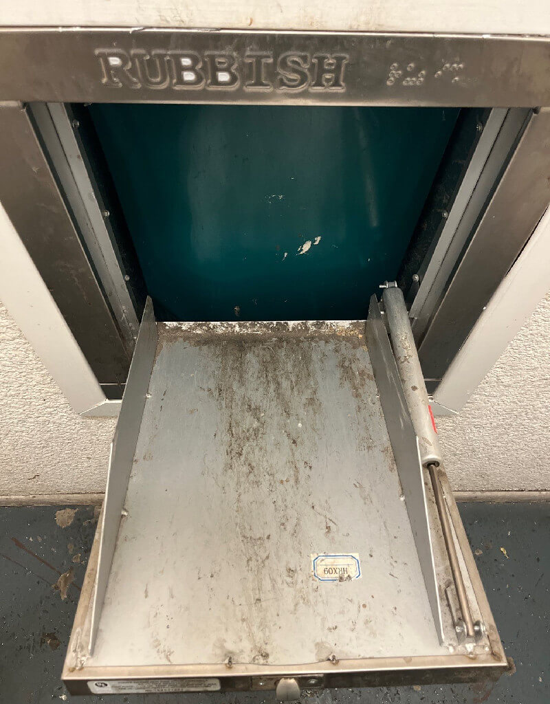 A dirty, metallic rubbish chute labeled "RUBBISH" is open, revealing a worn, angled tray with grime and debris. The chute door is blue inside, surrounded by a stainless steel frame, attached to a white wall. The chute looks old and used.