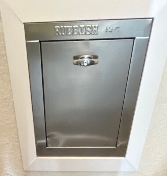 A small metal rubbish chute door set in a white frame. The door has a handle and "RUBBISH" engraved at the top, along with faint additional markings. The surface is shiny and slightly reflective.
